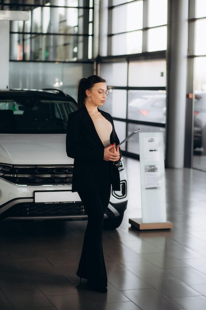 pretty young saleswoman in elegant suit dealer standing near car at new modern dealership