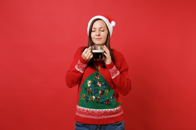 Pretty young Santa girl in Christmas hat holding, sniffing cup of coffee keeping eyes closed isolated on red wall background. Happy New Year 2019 celebration holiday party concept. Mock up copy space.