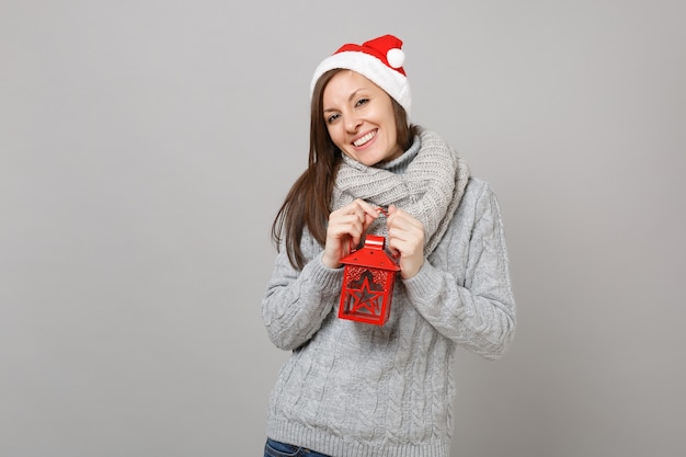 Pretty young Santa girl in gray sweater, scarf Christmas hat holding vintage lantern candlestick isolated on grey background. Happy New Year 2019 celebration holiday party concept. Mock up copy space.