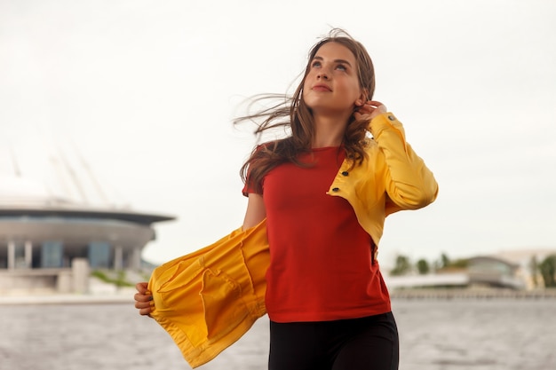 Photo pretty young sexy woman of athletic build stands against background of sea and building back. walk through the city center. portrait of cute female in yellow jacket and black trousers on street