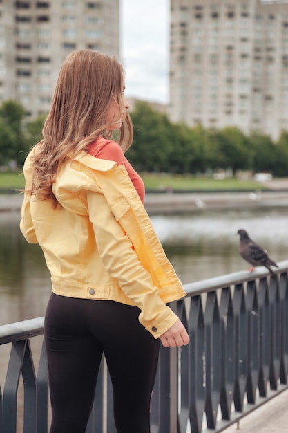 Pretty young sexy woman of athletic figure in bright clothes walking on canal embankment in city. Walk in city center. Portrait of cute pretty female in yellow jacket and black pants on street