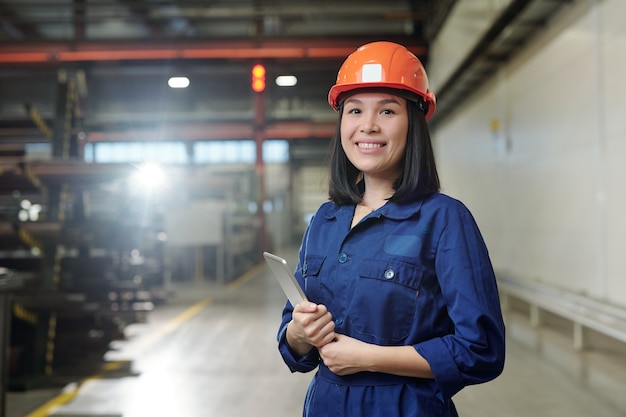 Pretty young smiling engineer in uniform and protective helmet holding tablet while posing