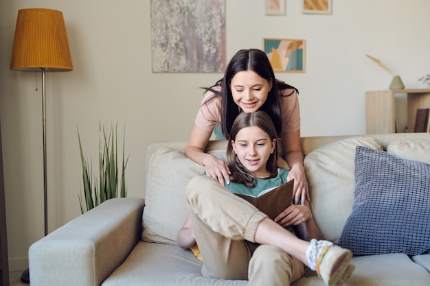 Pretty young smiling female with her chin on head of clever teenage daughter with open book reading together while staying home