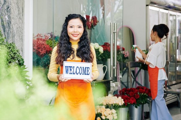 Pretty young smiling flower shop owner welcomming customers inside, her colleague spraying flowers