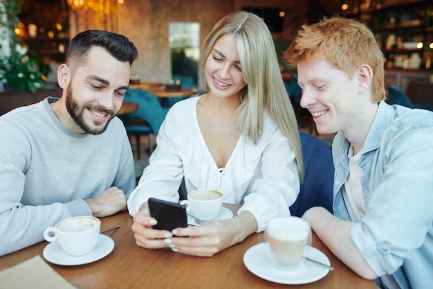 Pretty young smiling woman and two happy guys watching video or images in smartphone while relaxing by cup of coffee in cafe