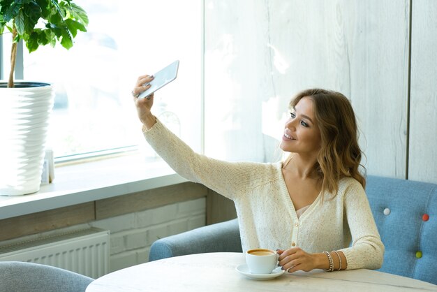 Pretty young smiling woman with smartphone making selfie while resting in cafe and having cup of coffee at leisure