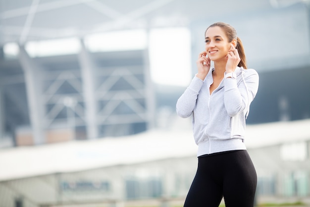 Pretty young sportswoman putting on headphones and listening to music in city