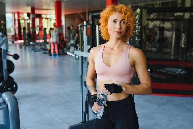 Pretty young sportswoman relaxing after training and drinking water in gym