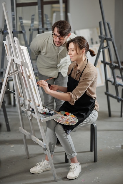 Pretty young student working over painting in front of easel while teacher bending over her during consultation at lesson