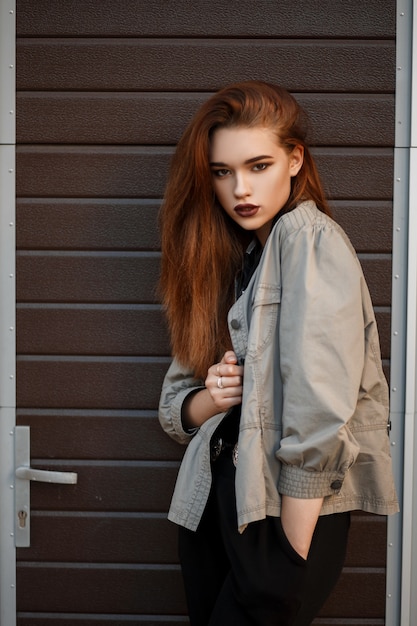 Pretty young stylish woman in a fashion spring jacket and black polo shirt stands near the gate on the street