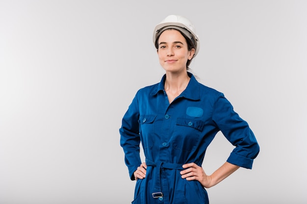 Photo pretty young successful female engineer in blue workwear and safety helmet keeping her hands on waist in isolation