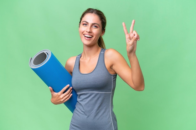 Pretty Young Uruguayan sport woman going to yoga classes while holding a mat over isolated background smiling and showing victory sign