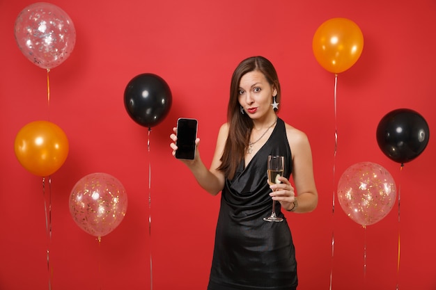 Довольно молодая женщина в черном платье, держа бокал шампанского, мобильный телефон с пустым черным пустым экраном на ярко-красном фоне воздушных шаров. С Новым годом, концепция праздничной вечеринки макет дня рождения.