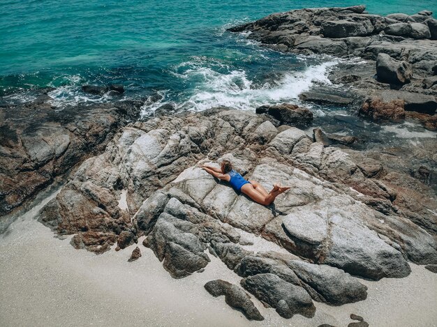 Pretty young woman in a blue swimming suit lying on the rocks in high tide. summer vacation concept