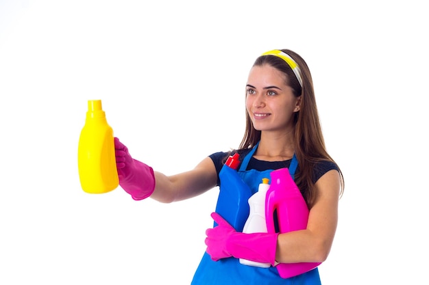 Pretty young woman in blue T-shirt and apron with pink gloves holding detergents on white background in studio