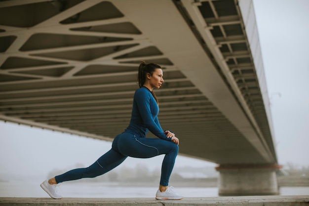 Piuttosto giovane donna in tuta blu che si estende prima dell'allenamento in riva al fiume al mattino d'autunno