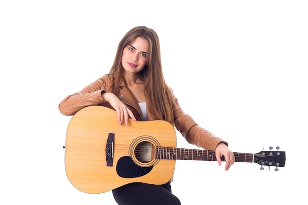 Pretty young woman in brown jacket and black trousers holding a guitar on white background in studio