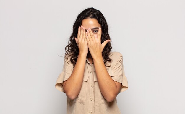 Pretty young woman covering face with hands, peeking between fingers with surprised expression and looking to the side