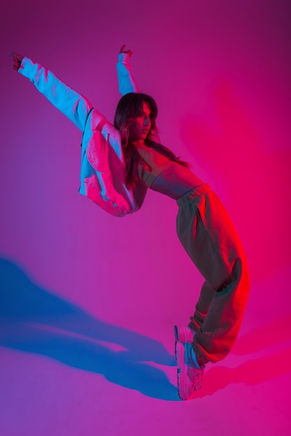 Pretty young woman dancer in fashionable youth clothes in a white sneakers dancing in a room with red light. Modern beautiful girl fashion model enjoys a dance in the studio with neon pink-blue color.