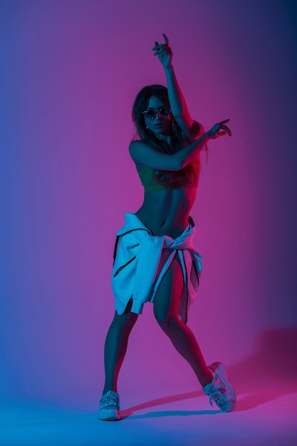 Pretty young woman dancer in a stylish top in a shoe in a hoodie dancing in the studio with a brightly neon blue light. Dance fashionable girl fashion model in sunglasses indoors with colored glitter