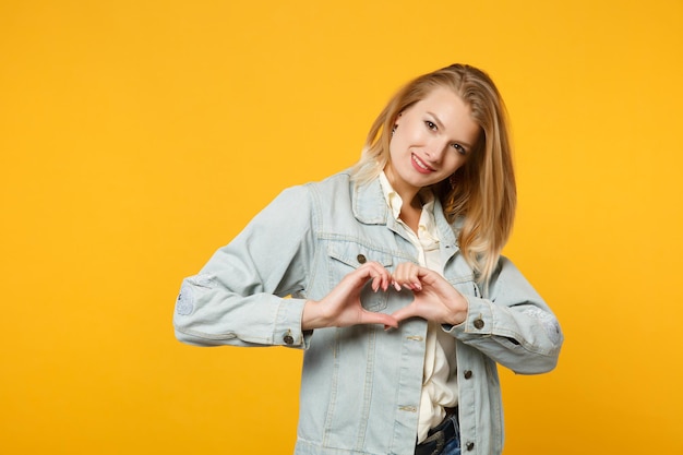 Piuttosto giovane donna in abiti casual denim guardando la fotocamera, mostrando il cuore di forma con le mani a forma di cuore segno isolato su sfondo giallo arancione in studio. concetto di stile di vita delle persone. mock up spazio di copia.