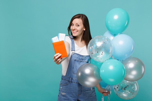 Pretty young woman in denim clothes holding passport boarding pass ticket celebrating with colorful air balloons isolated on blue turquoise background. Birthday holiday party, people emotions concept.