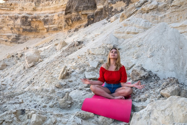 Pretty young woman doing relax yoga exercise with mat on dry sand among empty quarry