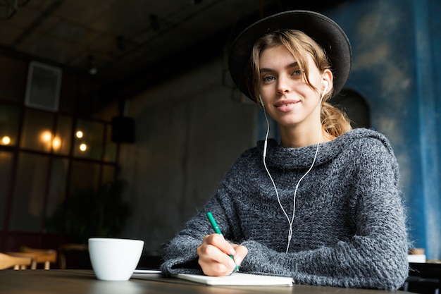 Pretty young woman dressed in sweater and hat sitting at the cafe table indoors, listening to music with earphones, drinking coffee, taking notes