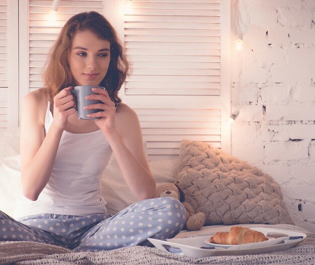 Pretty young woman drinking coffee sitting on bed at home