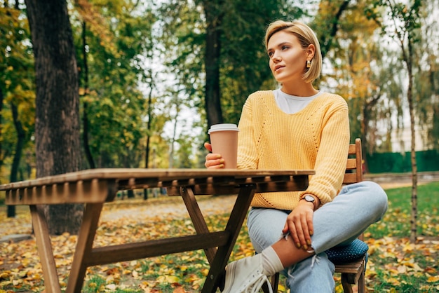 Pretty young woman drinking coffee at the terrace cafeteria in the park Blonde female in the city street drinking a hot beverage looking at one side Pretty girl with a cup of coffee on the go