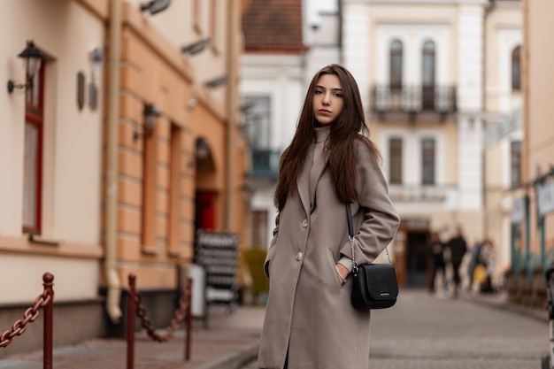 Симпатичная молодая женщина в элегантном пальто со стильной черной сумочкой стоит на улице в городе