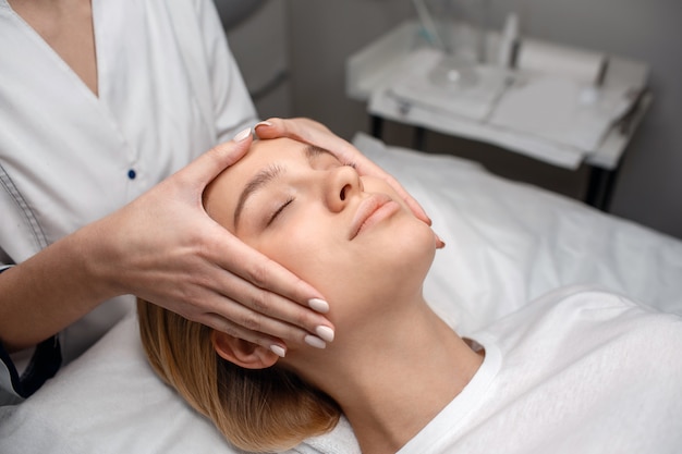 Pretty young woman enjoy face massage on couch. Beautisian touch it with bare hands. She stand at her head.