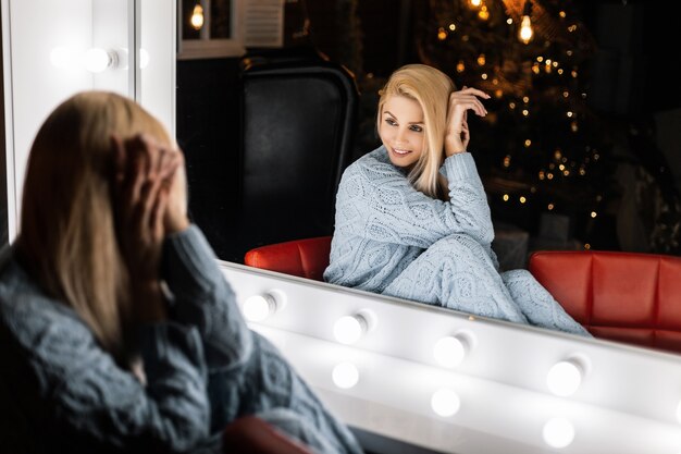 Pretty young woman in a fashion knitted suit is sitting near a vintage mirror with bright lamps in the living room in the evening time