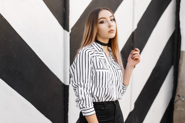 Bella giovane donna in una camicia bianca e nera alla moda in una linea con un naso forato in jeans neri in una collana di velluto in posa vicino a un muro vintage a strisce