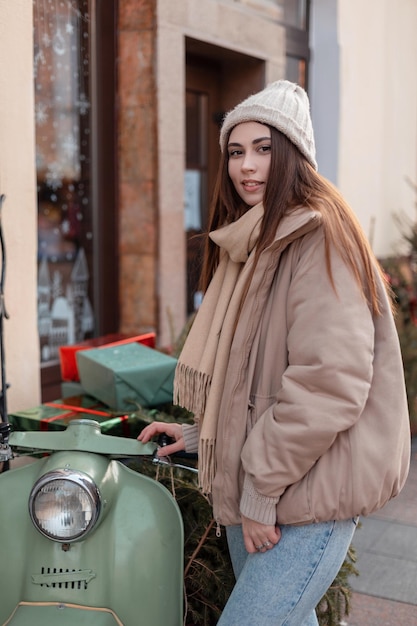 Bella giovane donna in un capospalla alla moda con un cappello lavorato a maglia, un'elegante giacca invernale e una sciarpa si trova vicino a una bici verde retrò con un albero di natale e regali