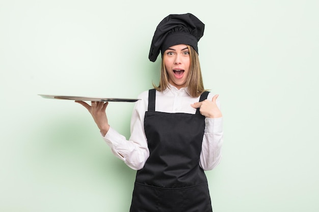 Pretty young woman feeling happy and pointing to self with an excited chef with a tray concept