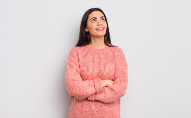 Pretty young woman feeling happy proud and hopeful wondering or thinking looking up to copy space with crossed arms