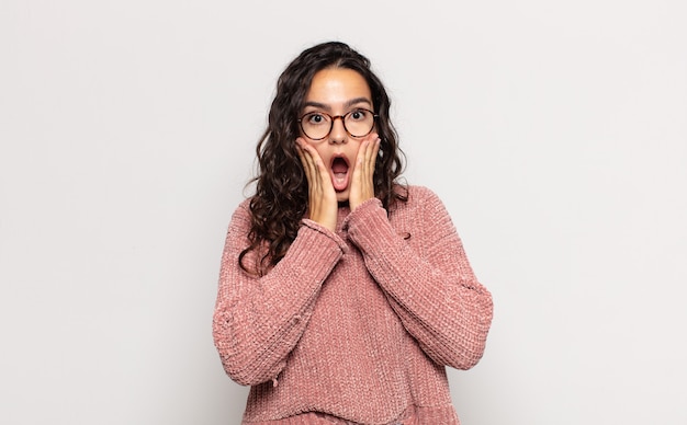 Pretty young woman feeling shocked and scared, looking terrified with open mouth and hands on cheeks