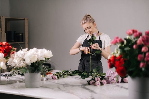 Pretty young woman florist wearing apron cutting fresh rose to bouquet at the table