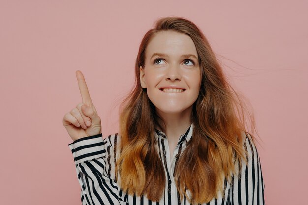 Photo pretty young woman in formal striped black and white blouse biting lower lip, demonstrating thinking process having pointer finger up posing against pink background. advertisement and promotion