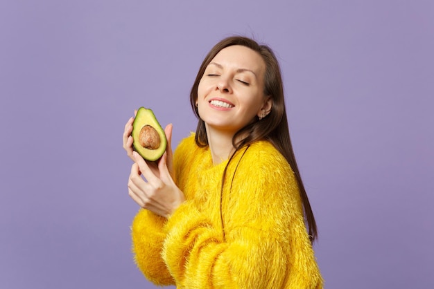 Pretty young woman in fur sweater keeping eyes closed holding half of fresh ripe green avocado isolated on violet pastel background. People vivid lifestyle relax vacation concept. Mock up copy space.