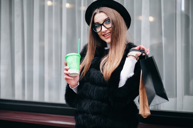 Довольно молодая женщина в очках гуляет по городу с чашкой кофе в руках после долгих покупок