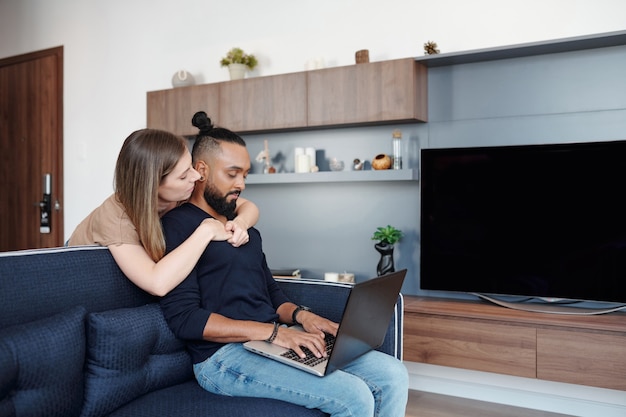 Pretty young woman hugging her boyfriend from behind when he is working on laptop when sitting on sofa at home