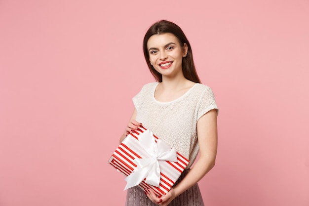 Pretty young woman in light clothes posing isolated on pink background. Valentine's Day Women's Day birthday, holiday concept. Mock up copy space. Holding red striped present box with gift ribbon bow.