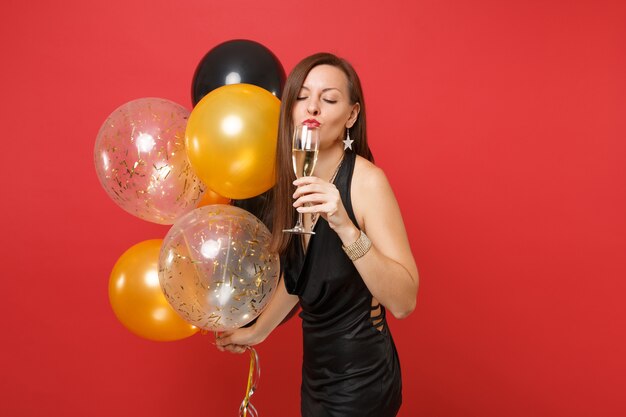 Pretty young woman in little black dress celebrating, drinking champagne, holding air balloons isolated on red background. St. Valentine's Day, Happy New Year, birthday mockup holiday party concept.