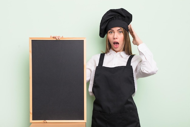 Pretty young woman looking happy, astonished and surprised. chef with blackboard