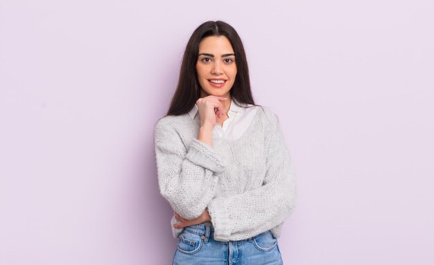 Pretty young woman looking happy and smiling with hand on chin, wondering or asking a question, comparing options