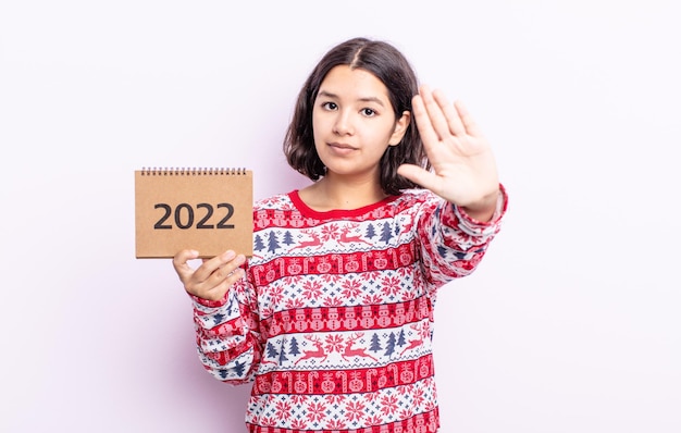 Pretty young woman looking serious showing open palm making stop gesture. 2022 calendar concept