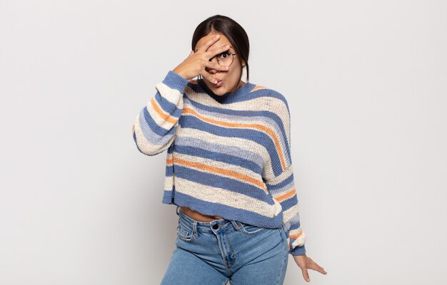 Pretty young woman looking shocked, scared or terrified, covering face with hand and peeking between fingers