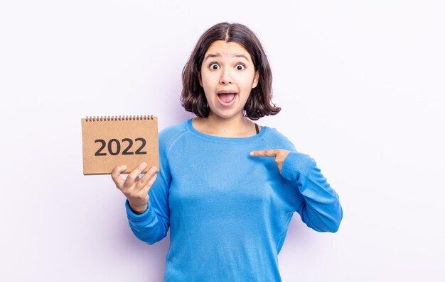 Pretty young woman looking shocked and surprised with mouth wide open, pointing to self. 2022 calendar concept